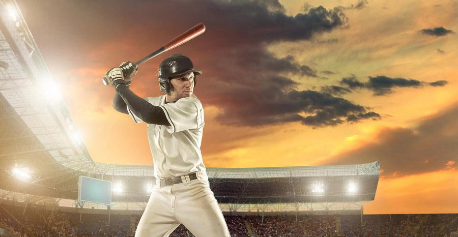 A modern bedroom showcases the Action Shot Wall Mural, depicting a baseball player swinging a bat at sunset. The room also has a bed, desk with computer, chair, window with blinds, photos on the wall, and a soccer ball on the wooden floor.