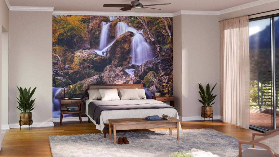 A bright bedroom with a bed, two nightstands, and potted plants features the stunning Autumn Waterfall Wall Mural. Sunlight streams through glass doors with sheer curtains onto the bench, rug, and serene decor.