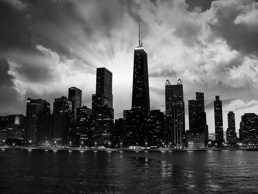 Product Description: The Chicago Skyline at Sunset Wall Mural captures the silhouette of a city skyline at dusk, featuring a prominent central skyscraper. Bright city lights outline the waterfront, casting vibrant reflections on the water. Dramatic clouds fill the sky, illuminated by bursts of light behind the tallest building. This stunning scene from would make an ideal custom printed mural for any space.