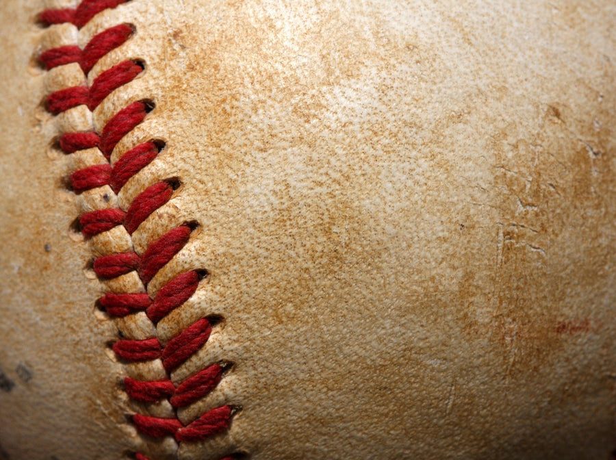A modern bedroom features the Baseball Macro Close Up Wall Mural with vivid red stitching. The space includes a bed, computer desk, chair, framed pictures, soccer ball under the bed, large white wardrobe, and sunlight through window blinds.