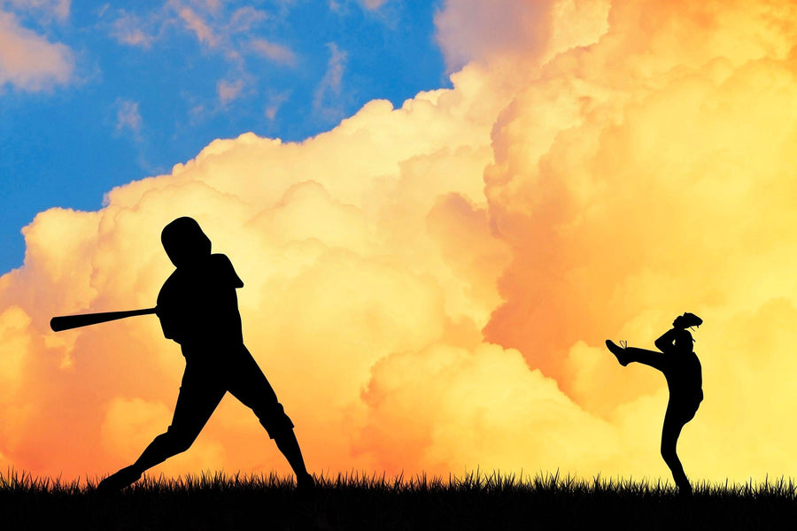 A bedroom with a workspace, bed, and the Baseball Silhouettes Wall Mural—durable peel and stick wallpaper showing two players (pitcher and batter) set against a bright orange-cloud sky. Sunlight streams through blinds; a ball lies near the bed.