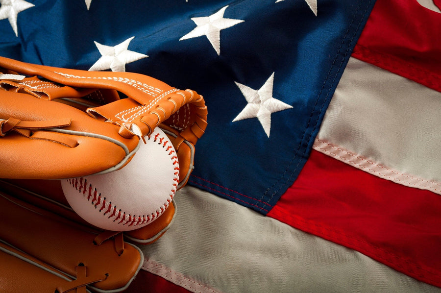 A bedroom showcases the Baseball and the Flag Wall Mural, depicting a glove holding a ball before an American flag. Sunlight highlights the desk, chair, bed, sports ball on the floor, framed pictures, and white cabinets.