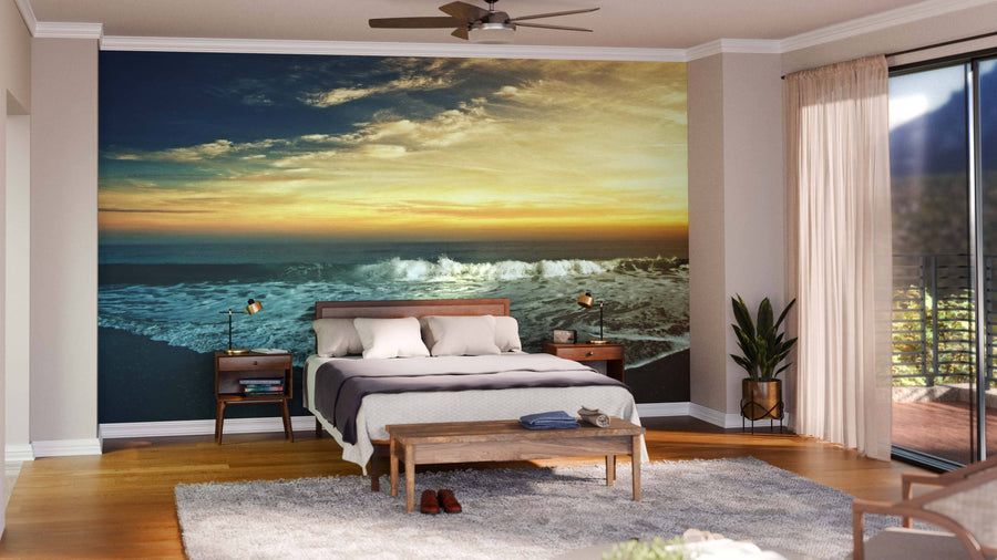 A modern bedroom features a wooden bed with gray bedding, matching nightstands, and a light area rug. Behind the bed is the Beach Panorama Wall Mural, depicting an ocean sunset. The room has hardwood floors, plants, and glass doors with sheer curtains.