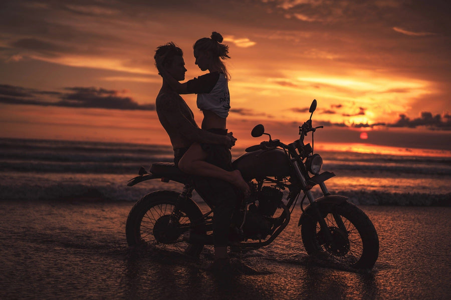 A tidy garage showcases dark cabinets and a vintage motorcycle. The back wall features the Beach Riders Wall Mural, showing two people silhouetted on a motorcycle at sunset by the beach. Tools are neatly arranged, sunlight comes in from the right.