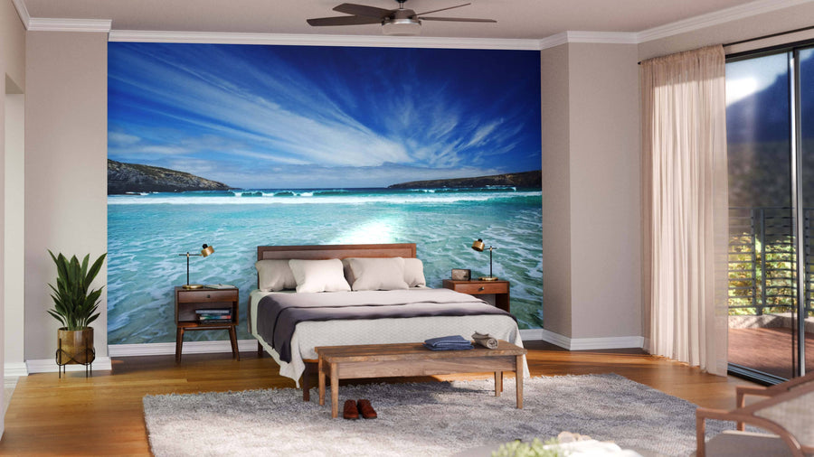 A modern bedroom showcases the Beautiful Bay Wall Mural, depicting a beach with blue sky and waves behind a wooden bed with white bedding. Nightstands, lamps, a plant, rug, bench, and large windows with beige curtains brighten the space.