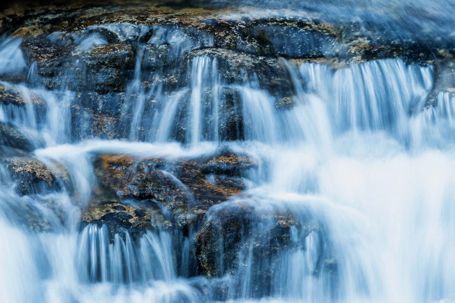 A modern bedroom features the Beautiful Waterfall Wall Mural behind the bed, wooden furniture, plants, a gray rug, and soft natural light streaming through sheer curtains and a glass door to the balcony. Light walls and a ceiling fan complete the space.