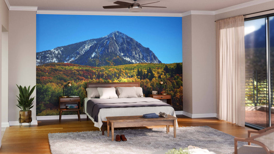 Modern living and dining room with wood flooring and mid-century furniture. The Beckwith Mountain Wall Mural adds vibrant color, covering one wall with a scenic mountain and autumn forest. Pendant lights hang above a dark dining table, chairs, and a rug.