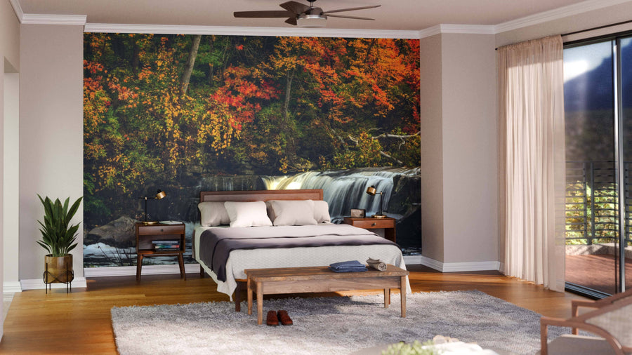 A modern open-plan living and dining room with wood floors, green chairs, a large dining table, and the Berea Falls In Autumn Wall Mural showcasing a waterfall amid vivid autumn trees. Warm hanging globe lights complete the inviting atmosphere.