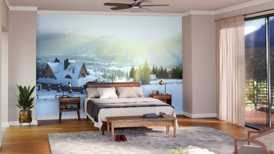 Modern living and dining room with wooden floors, stylish furniture, and a Big Winter House Mountain Wall Mural depicting a snowy village at sunrise. Blue and wooden chairs, a patterned rug, and a dark dining table under round pendant lights complete the space.