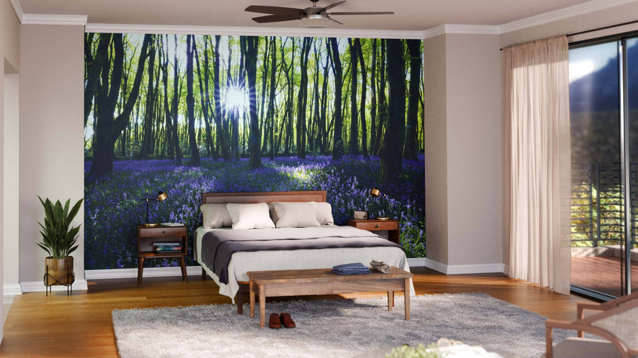 Modern living-dining area with wooden floors, teal armchairs, gray sofa, dining table for six. A Bluebell Wood Forest Wall Mural shows a sunlit forest with green trees and purple flowers. Pendant lights hang above the table; kitchen behind.