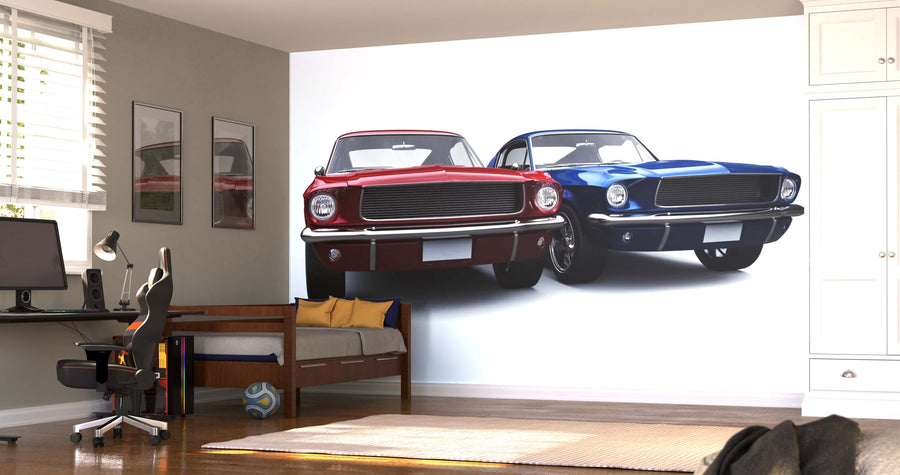 A modern bedroom with a bed, desk, and chair showcases the American Muscle Cars Wall Mural featuring two classic cars—one red and one blue—alongside wooden floors, framed car photos, and sunlight streaming through white blinds.