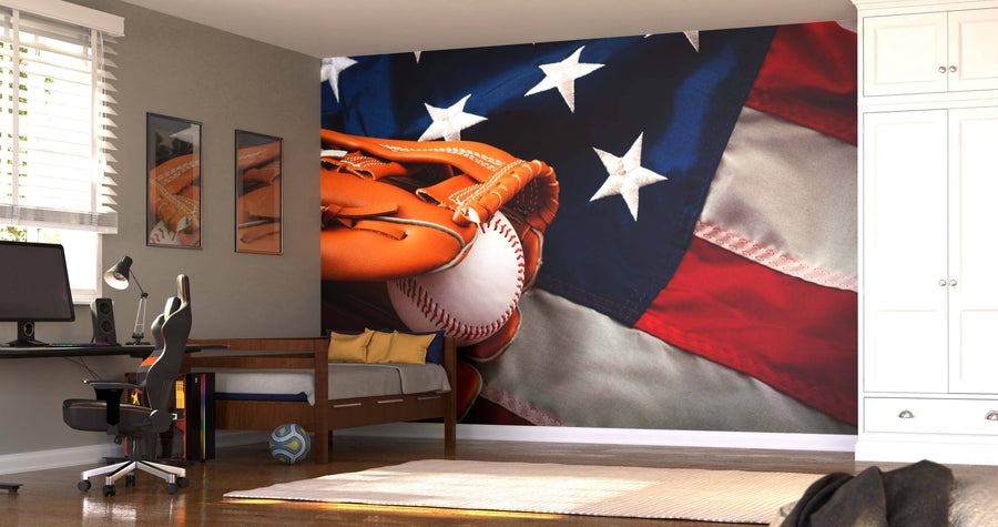 A bedroom showcases the Baseball and the Flag Wall Mural, depicting a glove holding a ball before an American flag. Sunlight highlights the desk, chair, bed, sports ball on the floor, framed pictures, and white cabinets.