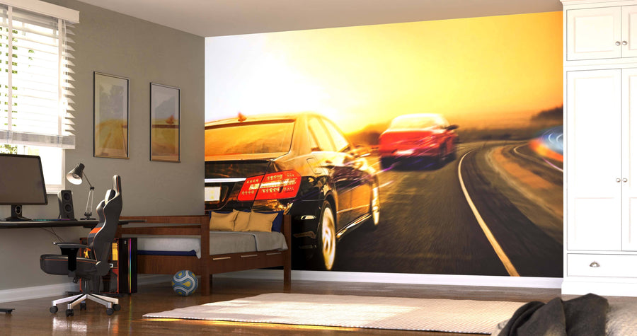 A tidy, sunlit bedroom with beige walls and modern furniture features the Cars Driving Fast Wall Mural, depicting two cars speeding on a highway at sunset. A desk, chair, bed, and sports ball complete the dynamic atmosphere.