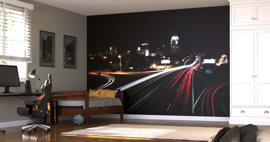 A modern bedroom with a desk, chair, twin bed, window with blinds, white wardrobe, and a soccer ball on the floor. The wall displays the Downtown Skyline and Passing Cars Wall Mural showing a city skyline at night with car light trails.