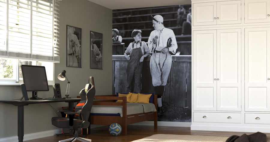 A bedroom features the Father and Son Standing Together Wall Mural, framed photos, a bed with yellow and navy pillows, a black desk with a computer and lamp, a gaming chair, a soccer ball on the floor, and large white cabinets.