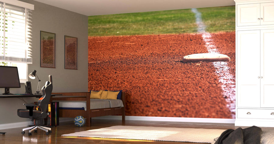 A bedroom with a desk, chair, bed, and wardrobe features the durable First Base Wall Mural—peel and stick, showing a close-up of a baseball base on reddish dirt with a white foul line. Light streams in through blinds on the window.