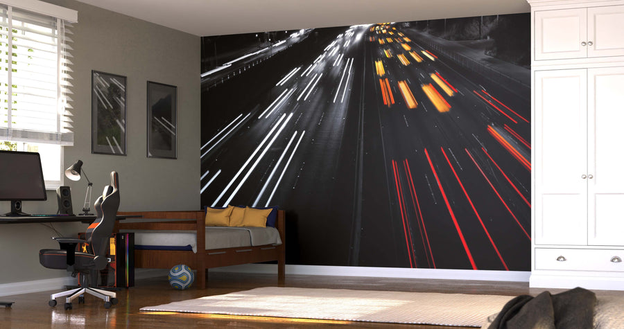 A modern bedroom with a desk, chair, and bed features the "Highway Traffic at Night Wall Mural," a peel-and-stick, removable wallpaper showing blurred white and red/yellow light trails of fast cars on a nighttime highway.