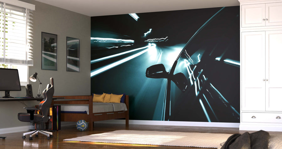 A modern bedroom with a desk, chair, and bed features the Night Drive Wall Mural, showcasing a blurred night highway scene. Sunlight streams through the window, illuminating the neutral space and adding dynamic movement.