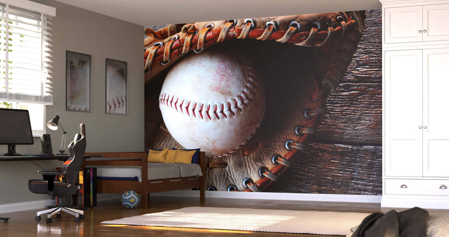 A modern bedroom with a bed, desk, computer, and chair features the Old Baseball Equipment Wall Mural—a close-up of a baseball in a worn glove on rustic wood. Sunlight shines through blinds, and a soccer ball rests near the bed.