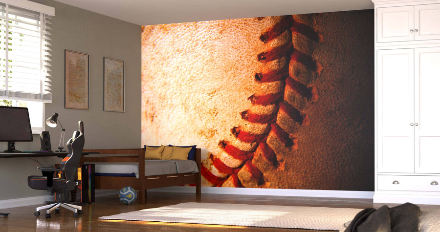 A modern bedroom features the Old Weathered Baseball Wall Mural with detailed red stitching, a single bed with navy and gold pillows, desk with computer, framed art, striped rug, soccer ball, white cabinets, and sunlight streaming in.