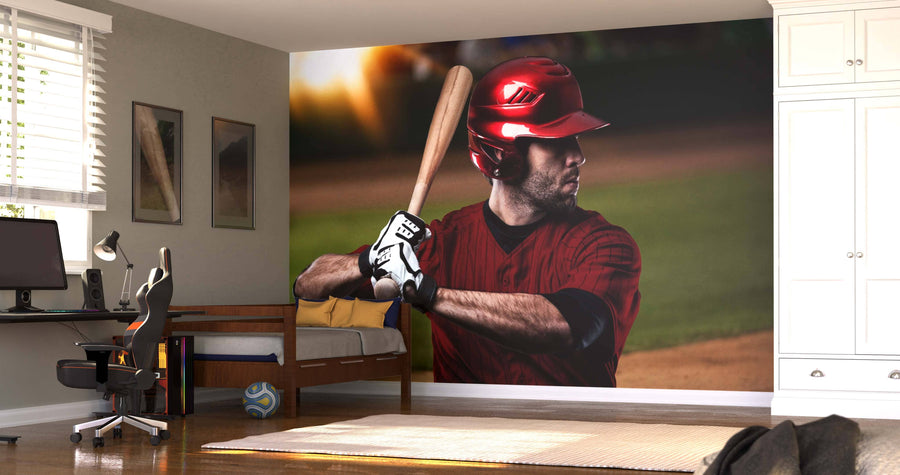 A bedroom with a bed, desk, and computer features the Power Stance Wall Mural of a baseball player in a red uniform mid-swing. Sunlight streams through the window, and a soccer ball rests near the bed on the wooden floor.