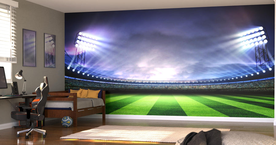 A bedroom decorated with the Stadium Glow Wall Mural—depicting a brightly lit soccer stadium at dusk—features a bed, computer desk, chair, soccer ball, and modern decor. Natural light through blinds enhances the sporty ambiance.