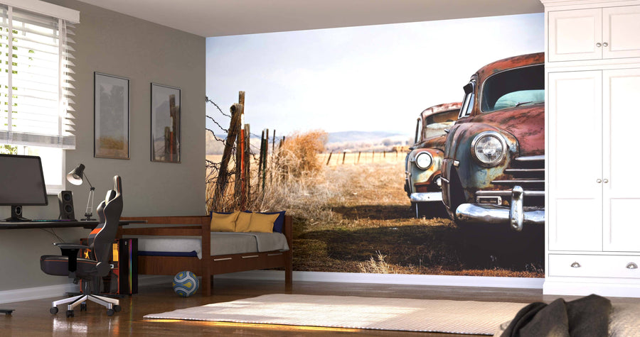 A bedroom with a workspace, bed, and wardrobe features the Vintage Cars Wall Mural showing two rusty vintage cars in a rural field. Sunlight streams in while a soccer ball lies on the wooden floor near the bed.
