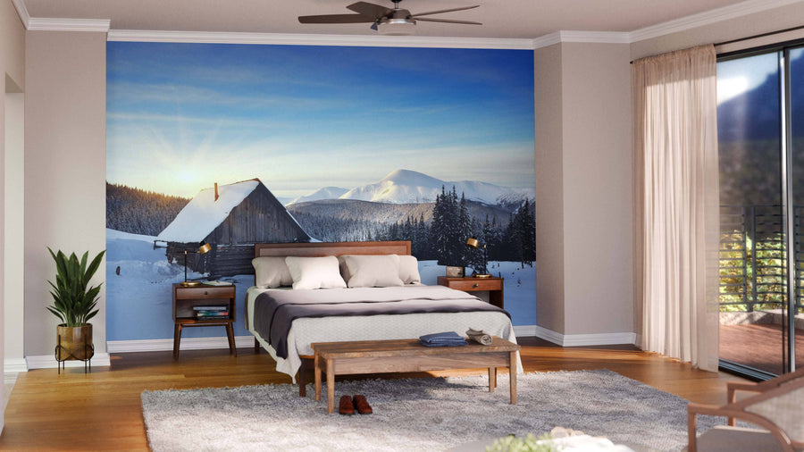 A modern living-dining room with wooden floors, teal armchairs, a rug, and a wooden dining table features the Cabin in the Mountains Wall Mural, adding a cozy and stylish window-like view of snowy mountains with cabins and sunrise.