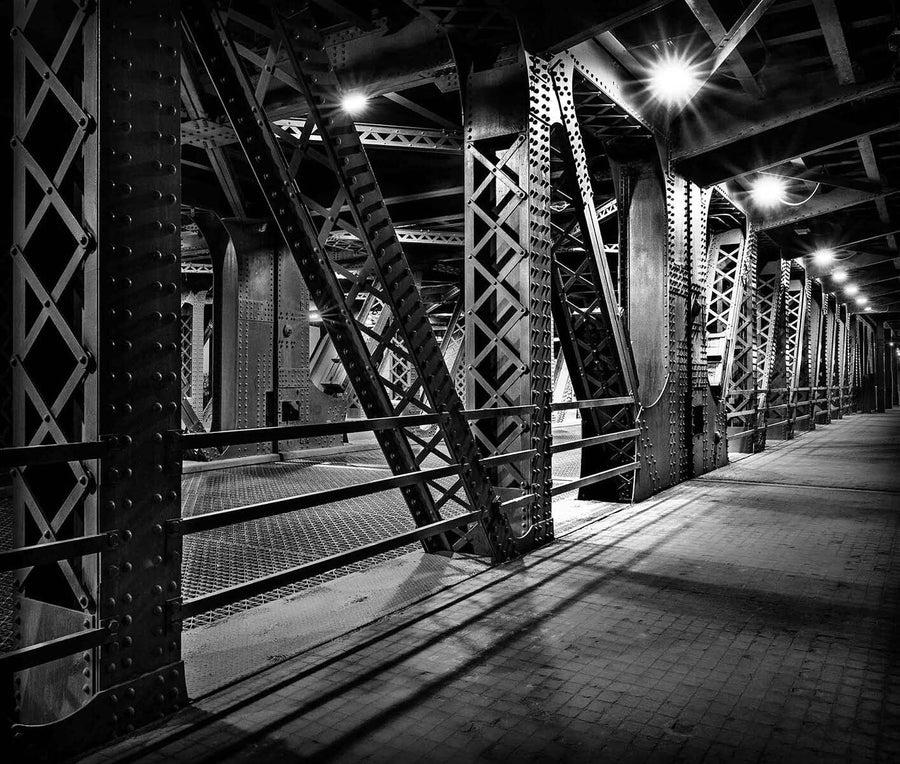 The Chicago Bridge Wall Mural depicts a dimly lit black and white scene of a metal bridge interior with crisscrossing beams, riveted columns, and geometric patterns that cast dramatic shadows on the concrete walkway, all softly illuminated by overhead lights.