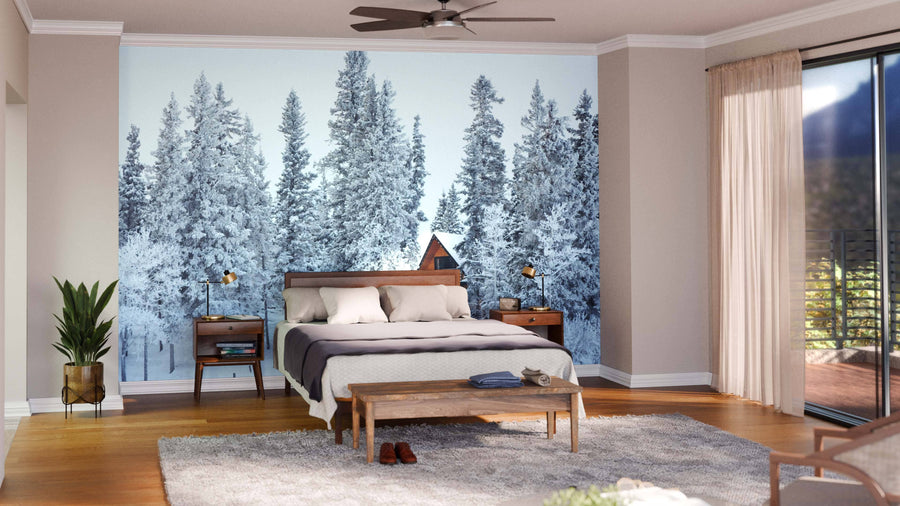 A cozy, modern living-dining room with wooden floors, dark green and wood furniture, large windows, pendant lighting above the dining table, and a wall-sized "Cottage in Winter Wonderland Wall Mural" depicting a snowy forest and cabin.
