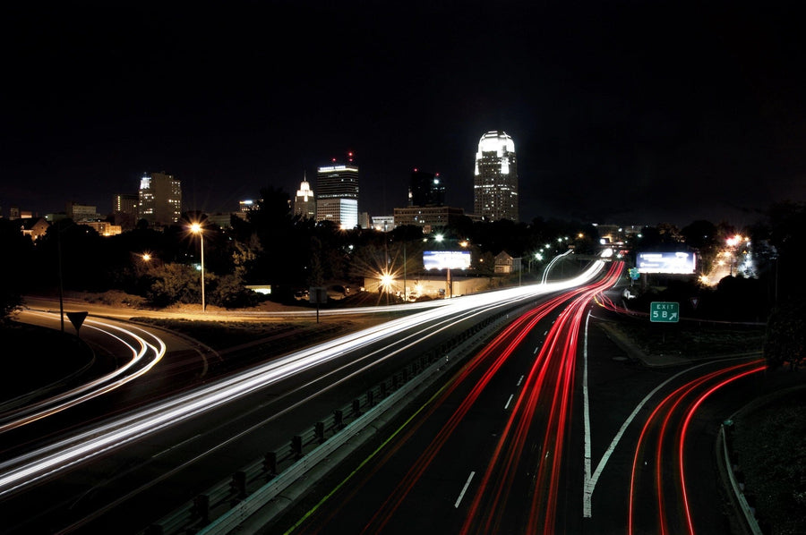A modern bedroom with a desk, chair, twin bed, window with blinds, white wardrobe, and a soccer ball on the floor. The wall displays the Downtown Skyline and Passing Cars Wall Mural showing a city skyline at night with car light trails.