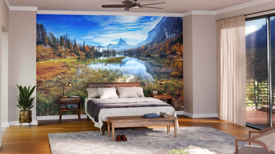 A modern bedroom showcases the Federa Lake Early Morning Wall Mural behind the bed, with neutral walls, white and gray bedding, wooden nightstands, a rug, plants, and sunlight streaming through sheer curtains on a sliding glass door.