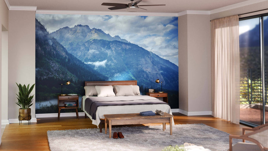 A modern bedroom features a wooden bed, two nightstands with lamps, a bench, a rug, and large windows. Behind the bed, the Fog in the Lowland Wall Mural adds dramatic flair and serves as removable wallpaper with majestic blue mountains.