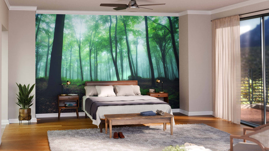 A modern bedroom features the "Foggy Morning in Green Forest Wall Mural" behind a wooden bed with white bedding and gray accents. Matching nightstands, a bench, potted plant, rug, and glass doors with beige curtains complete the tranquil space.