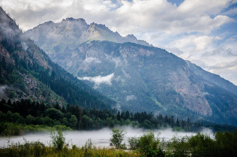 A modern bedroom features a wooden bed, two nightstands with lamps, a bench, a rug, and large windows. Behind the bed, the Fog in the Lowland Wall Mural adds dramatic flair and serves as removable wallpaper with majestic blue mountains.
