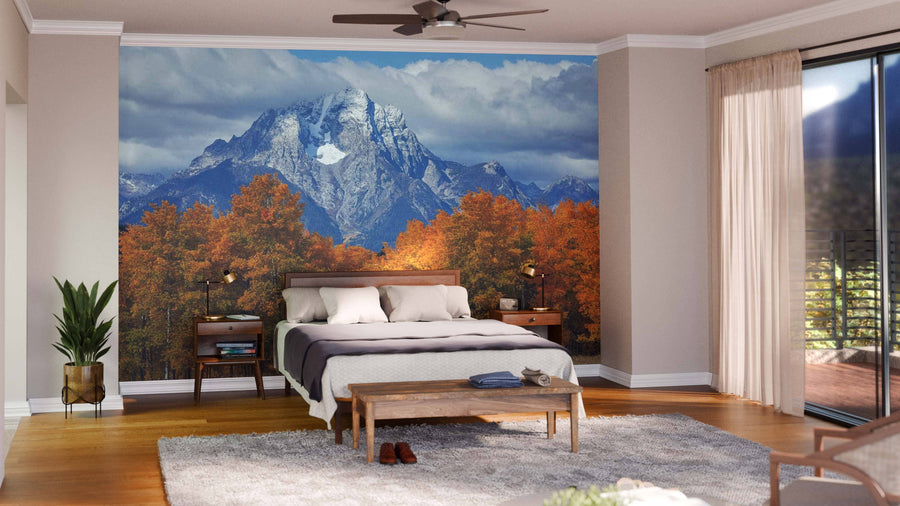 A modern open-plan living and dining room with wooden floors, mid-century furniture, and a Grand Teton Mountains Wall Mural depicting snow-capped peaks above autumn trees. Pendant lights hang over a dark dining table; a cozy seating area sits to the left.