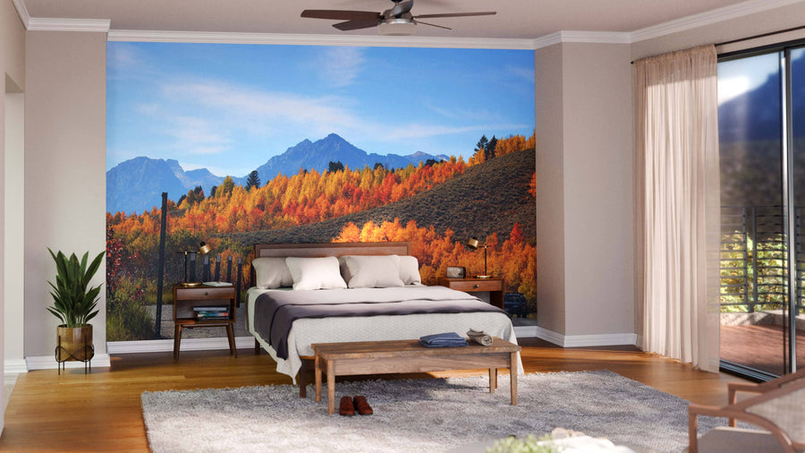 A modern living and dining room with wood floors, green chairs, a wooden table set, and the Grand Teton Mountain Range Wall Mural showcasing an autumn mountain landscape with a blue sky and vibrant red, orange, and green trees.