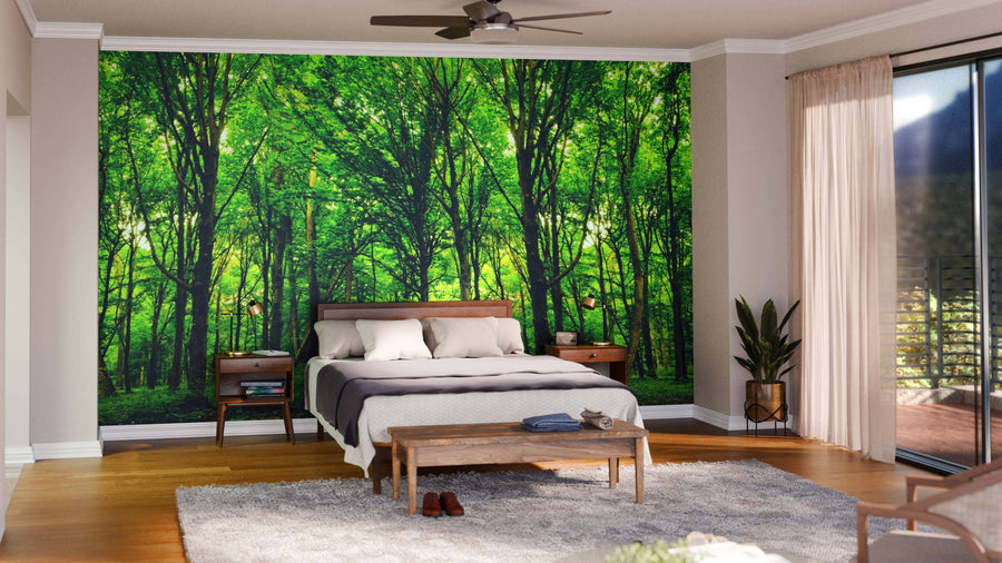 A bedroom features the Green Spring Forest Trees Wall Mural behind a wooden bed with white and gray bedding. Two nightstands with lamps, a wooden bench, gray rug, and a potted plant by sheer curtains complete the serene look.