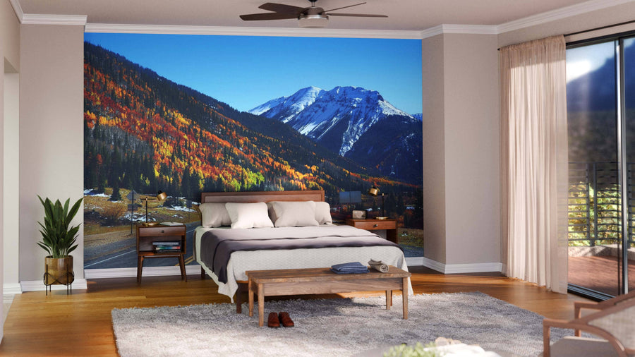 A modern living and dining room with wood floors, contemporary furniture, and globe pendant lights is highlighted by the "Highway to San Juan Mountains Wall Mural," depicting a mountain road through vibrant autumn trees and snowy peaks under a blue sky.