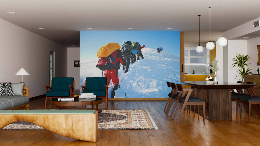 A modern living-dining area with wooden furniture and mid-century decor features the "Hike in Winter Mountain Wall Mural," displaying climbers ascending a snowy peak and infusing adventure into the cozy indoor setting.