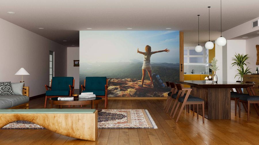A modern living-dining area with wood floors, teal chairs, and pendant lights features the Woman Backpacker Wall Mural—a peel and stick mural of a woman on a mountaintop, arms outstretched, gazing over a scenic valley at sunset.