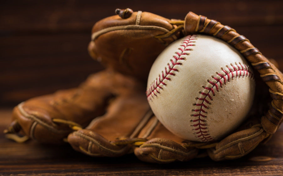 A bedroom with a desk, chair, and bed features the Leather Baseball Glove and Ball Wall Mural showing a close-up of a baseball in a glove. Sunlight streams in, and a blue and white soccer ball rests on the wooden floor by the bed.