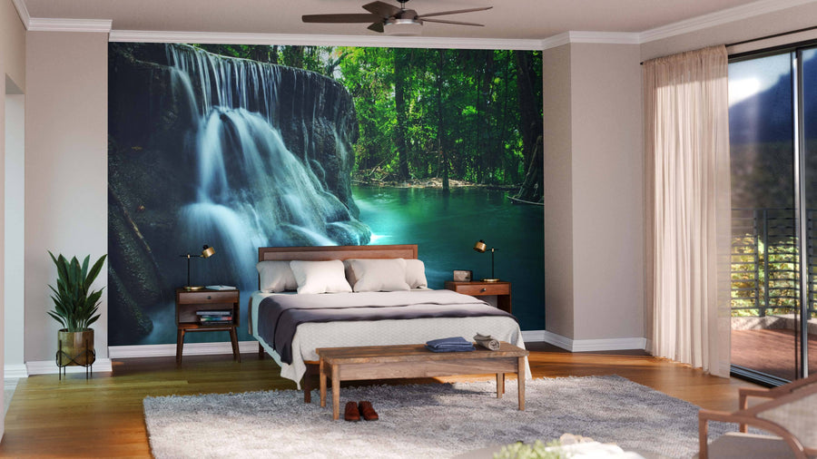 A bright, open living and dining area with wooden floors, teal and brown furniture, a patterned rug, and the Lime Stone Waterfall Wall Mural showcasing a lush green forest. Pendant lights illuminate the dining table.