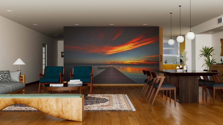 A modern living-dining room with wooden floors and mid-century furniture features the Beautiful Sunset Above the Sea Wall Mural as a bold focal point. Warm lighting, rugs, and stylish decor complete the cozy, contemporary vibe.