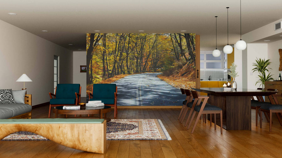 A modern living and dining room with wooden floors features the Birch Tree Highway Wall Mural, depicting a sunlit forest road with autumn trees. The space includes teal chairs, a gray sofa, wooden bench, dining table, and globe lights.
