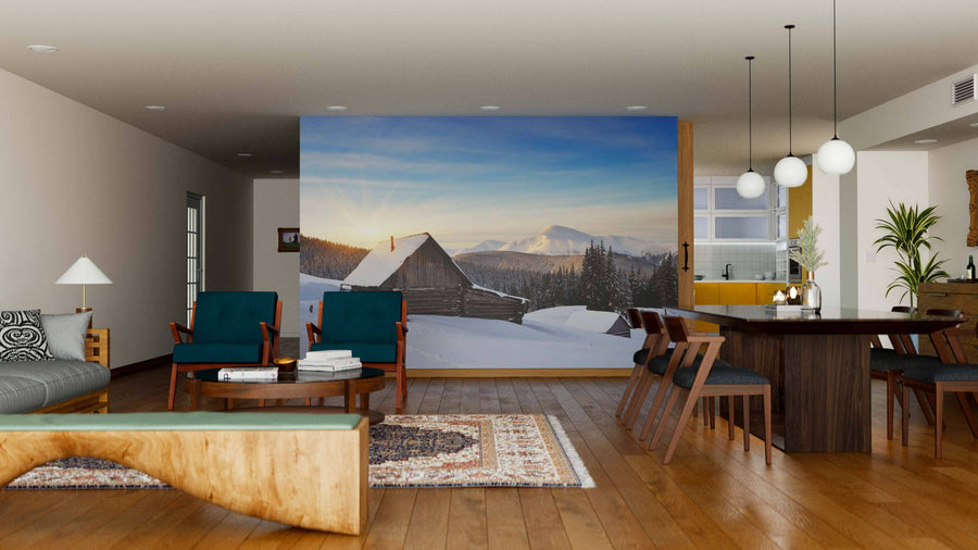 A modern living-dining room with wooden floors, teal armchairs, a rug, and a wooden dining table features the Cabin in the Mountains Wall Mural, adding a cozy and stylish window-like view of snowy mountains with cabins and sunrise.