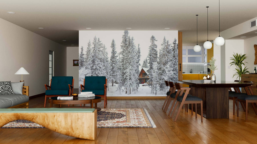 A cozy, modern living-dining room with wooden floors, dark green and wood furniture, large windows, pendant lighting above the dining table, and a wall-sized "Cottage in Winter Wonderland Wall Mural" depicting a snowy forest and cabin.
