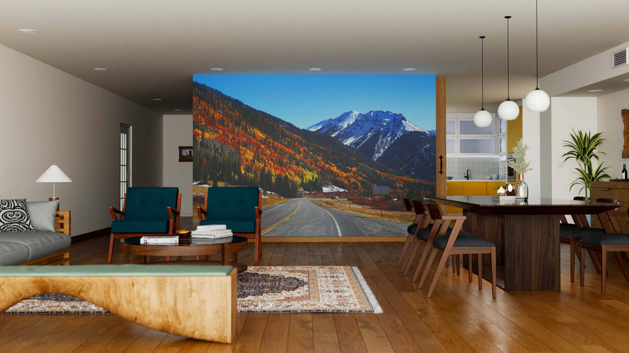 A modern living and dining room with wood floors, contemporary furniture, and globe pendant lights is highlighted by the "Highway to San Juan Mountains Wall Mural," depicting a mountain road through vibrant autumn trees and snowy peaks under a blue sky.