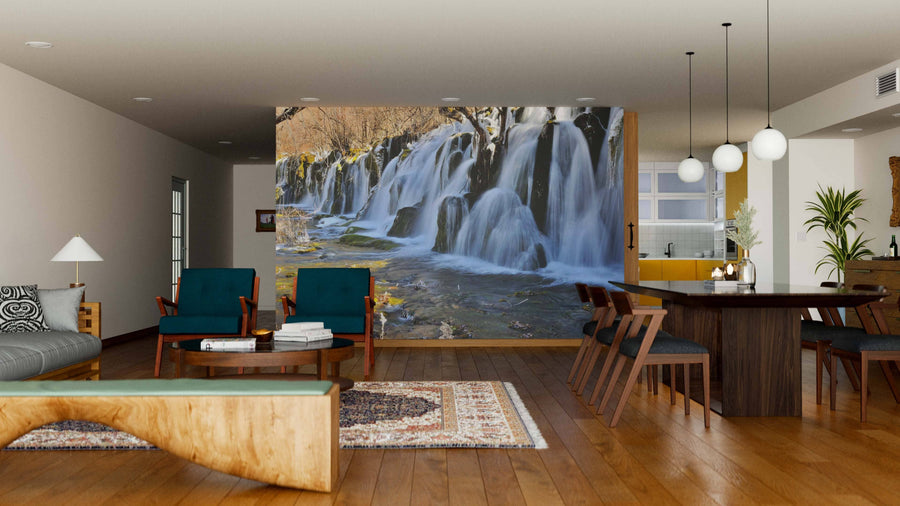 A modern living-dining room with wood floors and mid-century furniture features a Jiuzhaigou Valley Waterfalls Wall Mural as a nature-inspired focal point. Warm lighting and earth tones enhance the cozy atmosphere.