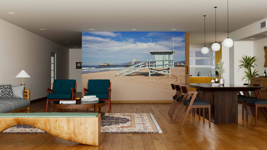 A modern living-dining room features wooden floors, mid-century furniture, and a Santa Monica Beach and Pier Wall Mural, creating an open coastal vibe. Pendant lights hang above the table and a rug accents the wood décor.